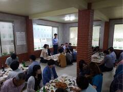 식당에서 음식이 놓여진 여러  테이블에 사람들이 둘러 앉아 있으며 한명이 서서 이야기를 하고 있고 앉아 있는 사람들이 경청하고 있는 모습 
